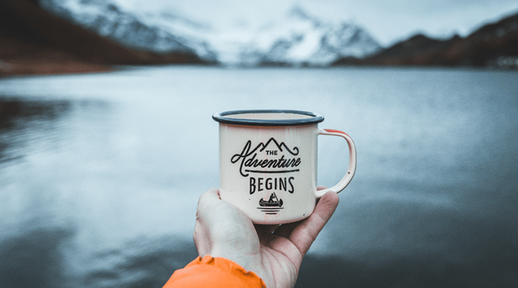 A hand with an orange sleeve by a lake and mountains holding an old fashioned white cup that says The Adventure Begins and shows a person rowing a canoe