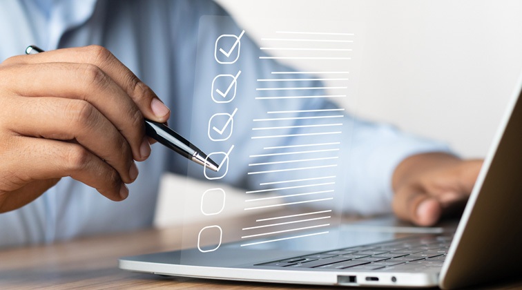 A close-up of a person in a long-sleeve shirt using a laptop and a pen crossing off a checkmark on a to-do list.