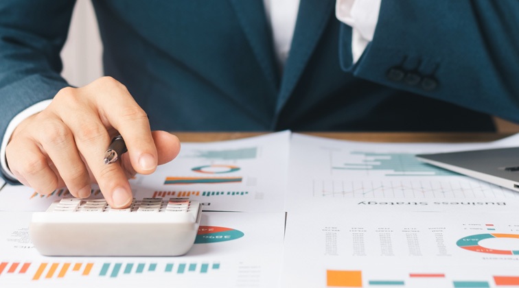 Close-up of a person in a suit with a pen and calculator at a desk looking at graphs and data on papers.