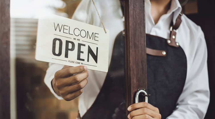 An employee wearing a white long-sleeve shirt with a black apron opening the store's front door and flipping their sign that says, Welcome We Are Open, Please Come In.
